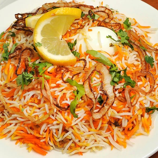 Colorful biryani rice dish with fried onions, cilantro, a hard-boiled egg, and lemon wedges.