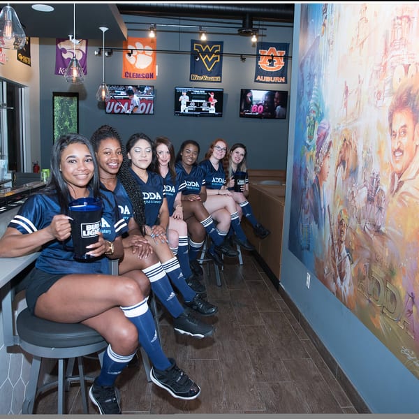 Six women in blue Adda Pub & Eatery uniforms sit at a bar with sports banners and a mural.