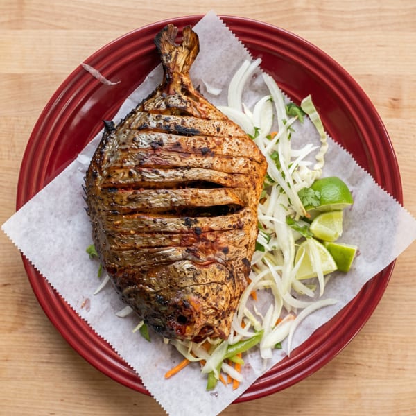 Grilled whole fish with crispy skin, served on a red plate with shredded cabbage, onions, and lime wedges.