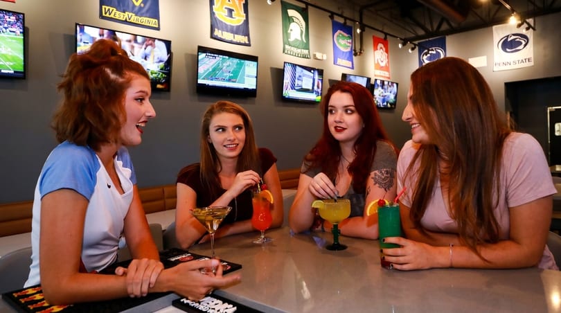 Four young women enjoying colorful cocktails at a bar with sports flags and TVs in the background.