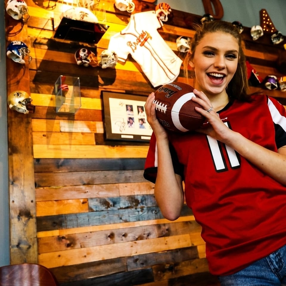 Smiling young woman in a red jersey holding a football in a sports-themed restaurant.