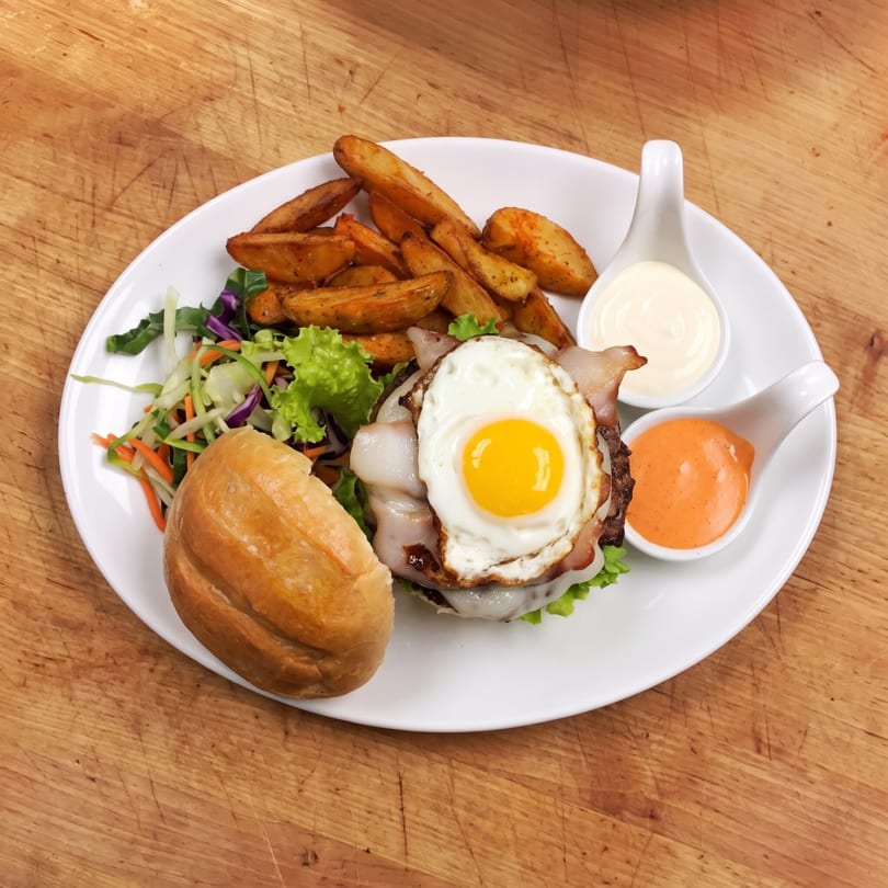 Gourmet burger with fried egg, bacon, cheese, potato wedges, salad, and two sauces on a white plate.