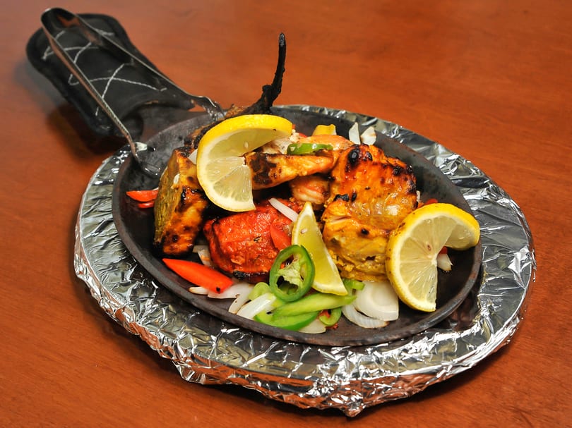 Mixed grill platter with chicken, shrimp, lemon, onions, and peppers on a sizzling cast iron pan.