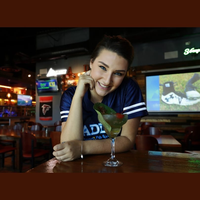 Smiling woman in a sports jersey with a cocktail at a bar, screens in the background.