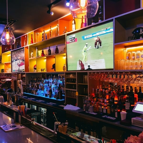 A vibrant bar interior with a long counter, liquor bottles, hanging glasses, and TVs showing sports.