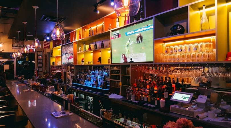 A vibrant bar interior with a long counter, liquor bottles, hanging glasses, and TVs showing sports.