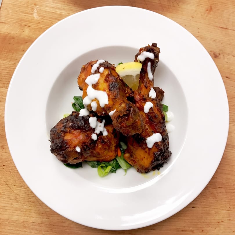 Four chicken drumsticks with white sauce and a lemon wedge on a bed of greens.