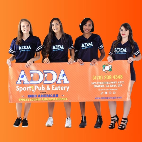 Four women in Adda Sports Pub & Eatery shirts holding a large orange banner with the restaurant's logo and details.