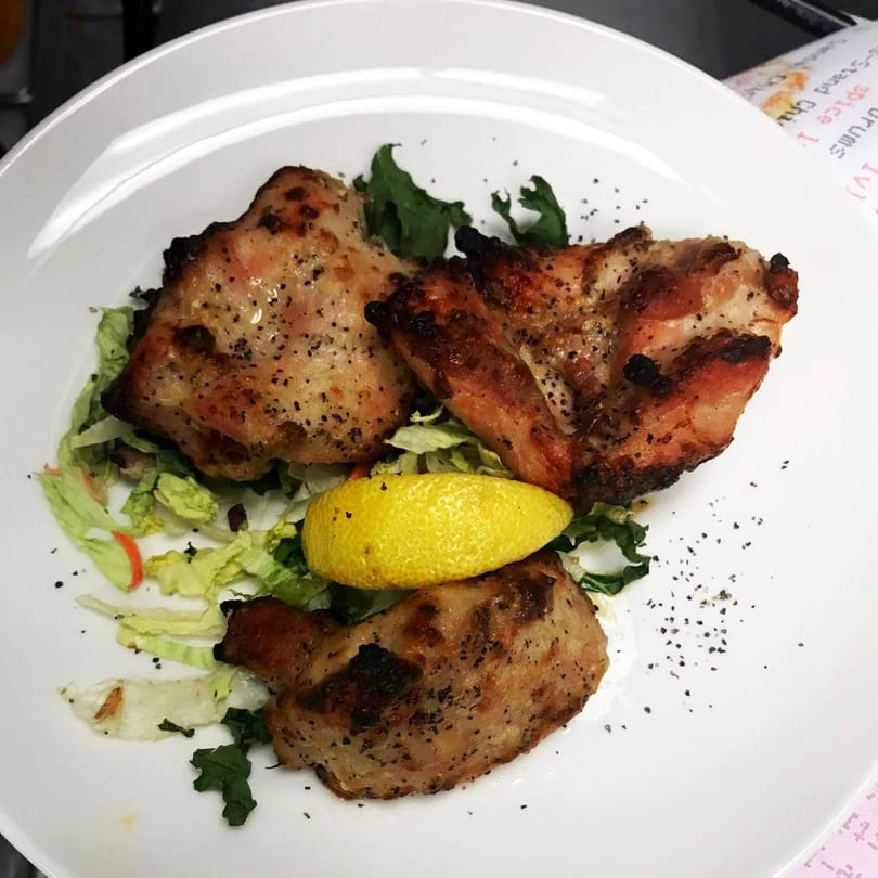 Three pieces of grilled chicken with black pepper, lettuce, and a lemon wedge on a white plate.